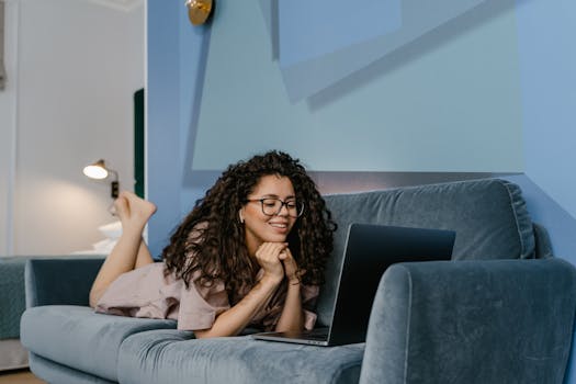 Happy woman with curly hair video calling on laptop, lying on sofa in cozy living room.